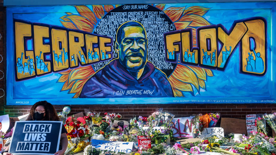 A mural of George Floyd with I CAN BREATHE NOW and many names behind him. Flowers, signs, and a person holding a BLACK LIVES MATTER sign are in front of the mural.