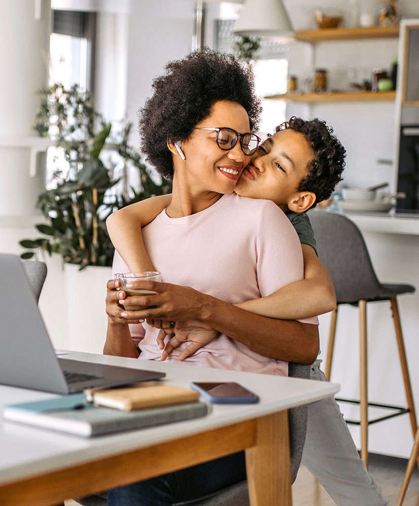 Working Mother at Home Taking a Break to Spend Time with Her Teenage Son. Boy is Hugging her from the Back.