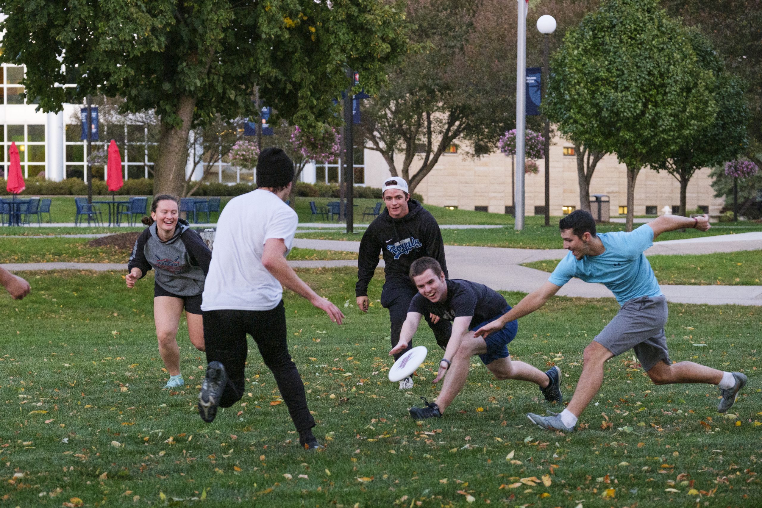 College students in a grassy area trying to catch a frisbee with trees and a building with windows in the background.