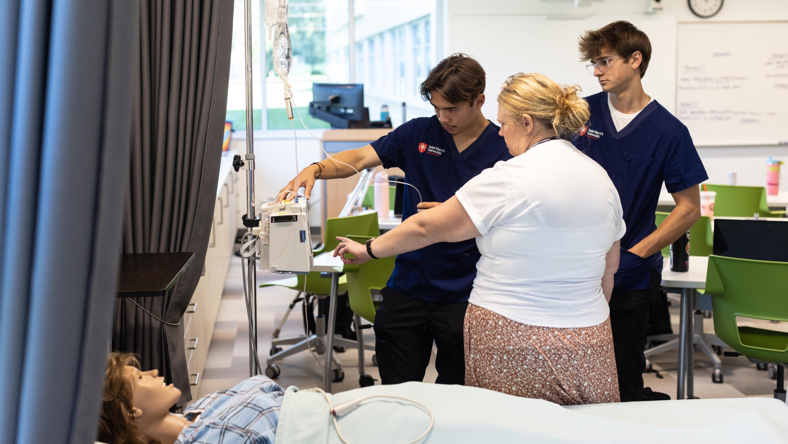 Two nursing students and an instructor adjusting an IV pump next to a medical training mannequin in a classroom.