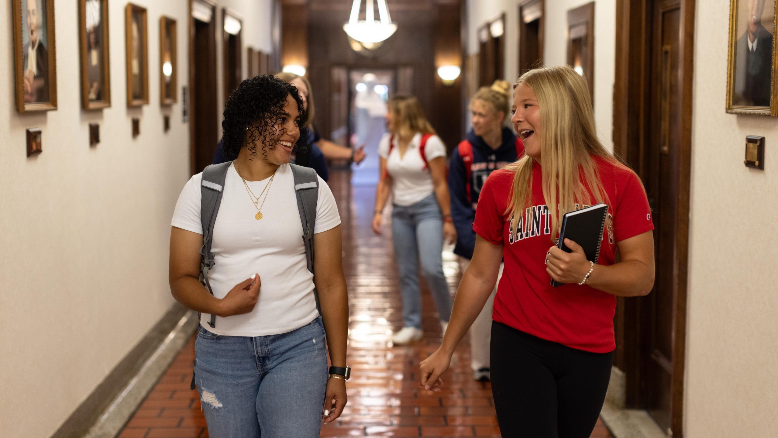 Two female students walk down a hallway lined with portraits, laughing as they talk to each other.