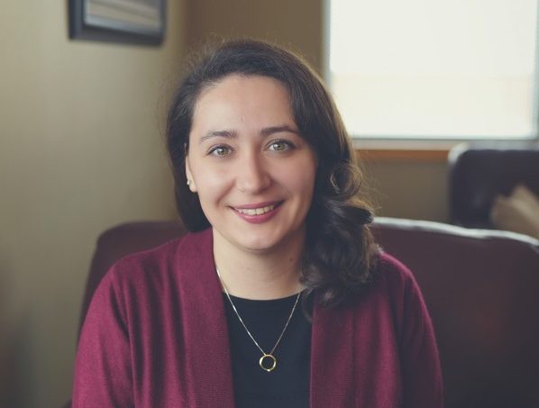 A smiling woman wearing a dark shirt, a burgundy cardigan, and a gold necklace sits in a chair.