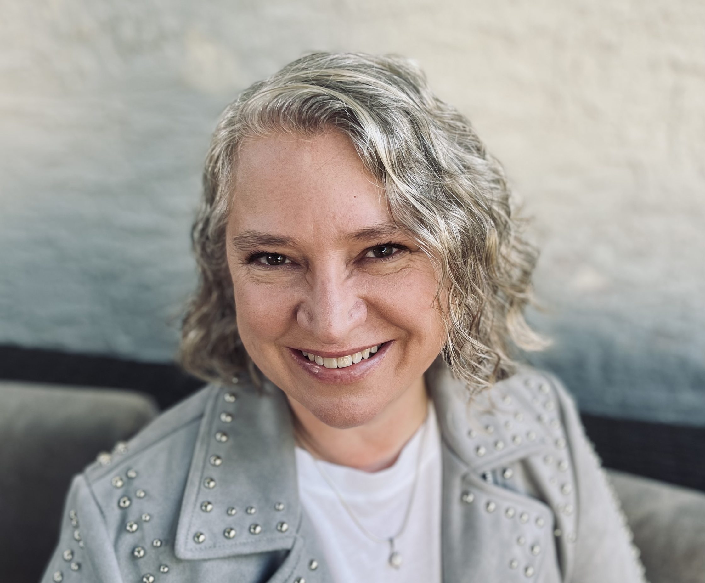 A smiling woman with wavy graying hair wearing a gray studded moto jacket over a white t-shirt.