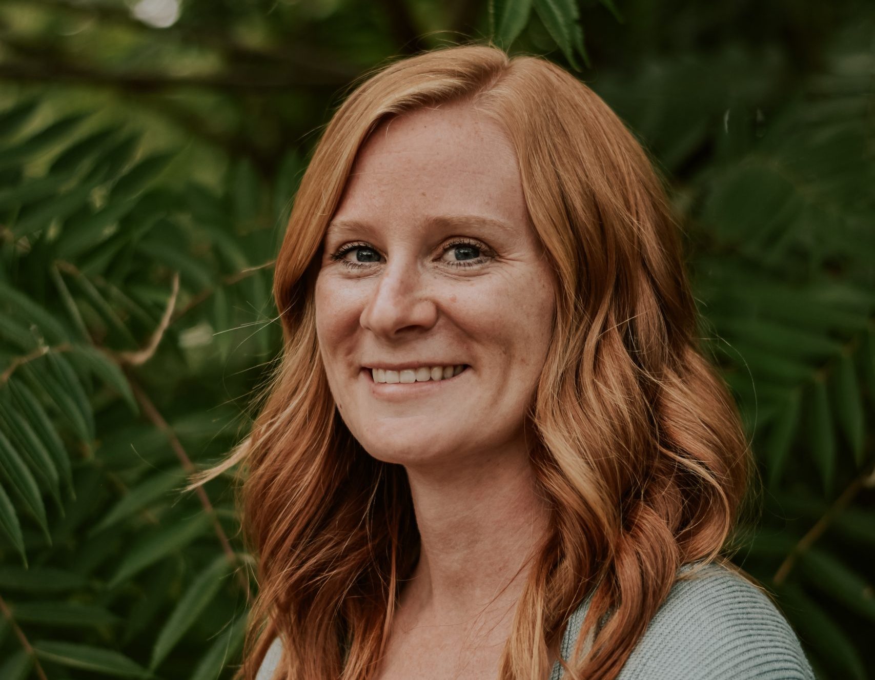 A smiling woman with long wavy red hair wearing a rust-colored top and a light blue cardigan stands in front of green leaves.