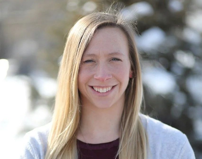 A woman with shoulder-length blonde hair smiles while wearing a maroon top and a grey cardigan.