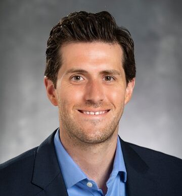 A man with short brown hair wearing a light blue button-down shirt and a navy blazer.
